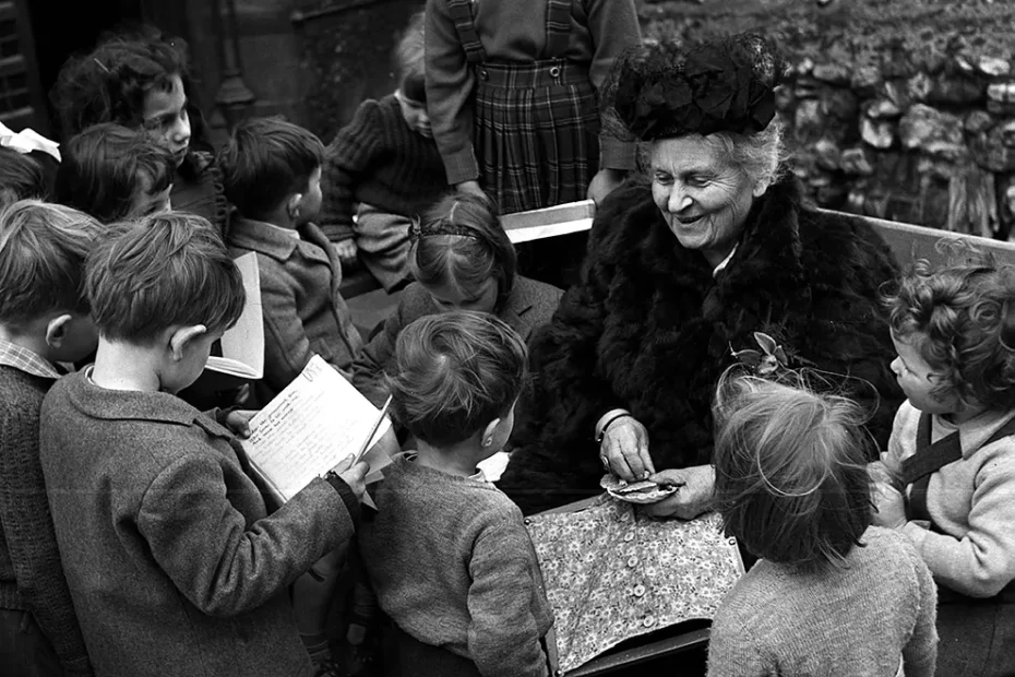 María Montessori interactuando con un grupo de niños en un entorno educativo, promoviendo el aprendizaje a través de la curiosidad y la participación activa.